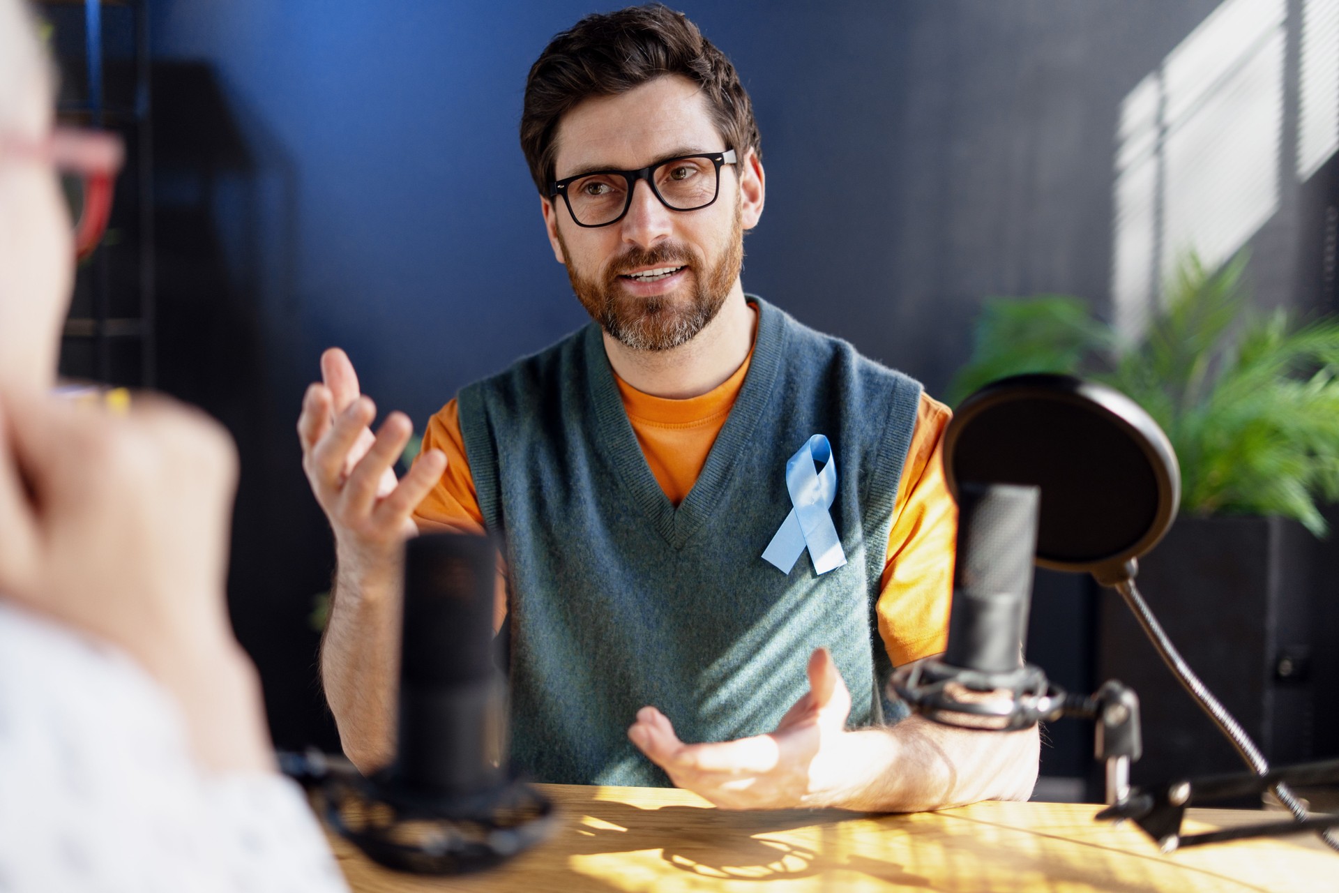 Radio host speaking with guest about prostate cancer awareness, wearing a light blue ribbon